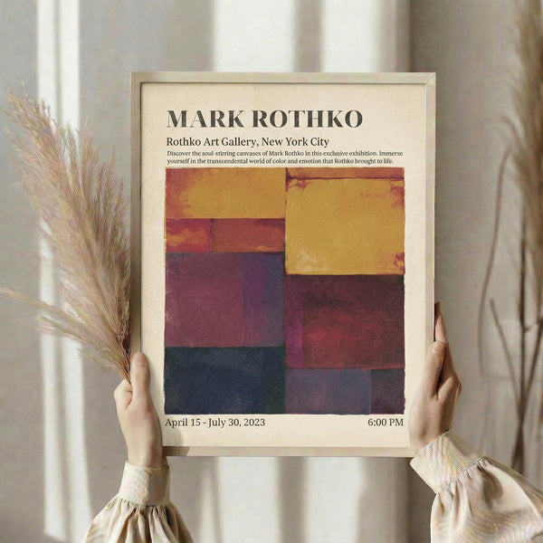 May include: Person holding framed exhibition poster featuring Mark Rothko artwork with geometric color blocks in warm yellows, deep reds, purples, and navy blue. Light wood frame against cream background with dried pampas grass. Person wearing beige knit sweater with gathered cuffs. Soft natural lighting creates gentle shadows on neutral wall.