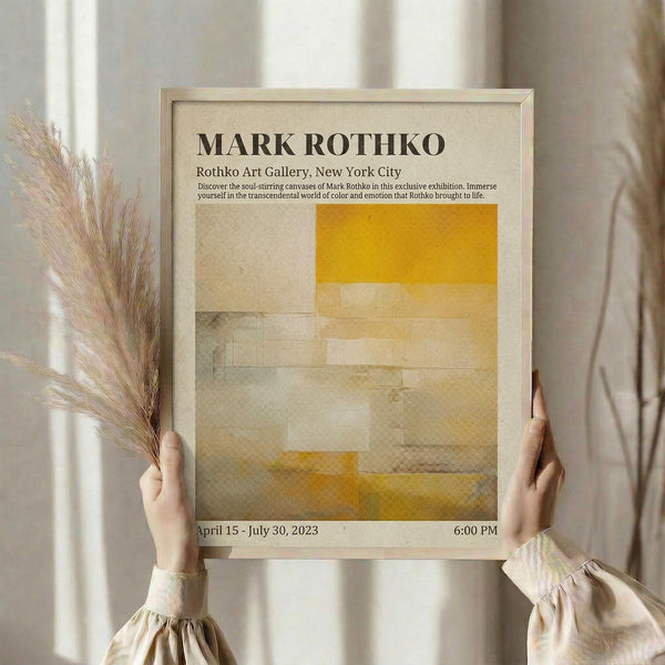 May include: Person holding framed exhibition poster featuring Mark Rothko artwork with yellow, beige and muted green abstract color blocks. Light wood frame against neutral curtained background. Dried pampas grass stems visible on left side. Soft natural lighting creates warm, minimalist aesthetic.