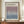 May include: Framed exhibition poster featuring Mark Rothko's name in bold text above a soft gradient square transitioning from dusty purple at top to blue-gray at bottom. The cream-colored poster shows gallery details and dates, displayed in a thin brown frame on a wooden sideboard next to a white ceramic vase and stacked books against white paneled walls.