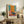 May include: Abstract art poster with orange, teal, and pink color blocks in the style of Mark Rothko, displayed in light wood frame on rustic wooden sideboard beside fern plant, ceramic vase, and stacked books in warm-toned living room setting with beige upholstered chair and natural lighting.