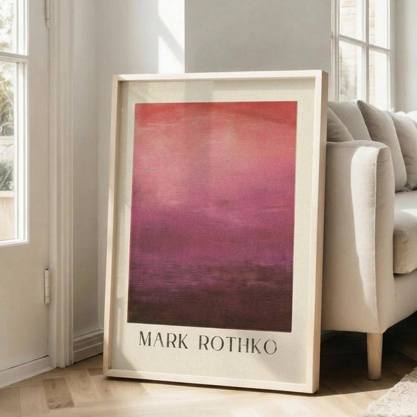 May include: A framed art print featuring a Mark Rothko-style abstract painting with horizontal bands of color transitioning from coral red at top through pink to deep burgundy at bottom. The print has a light wood frame with white matting and the artist's name printed below. It leans against a white wall next to a beige upholstered sofa in a bright room with tall windows and natural light casting soft shadows on hardwood floors.
