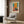 May include: Abstract art poster with orange and teal color blocks in warm, painterly style hanging on wood-paneled wall above beige upholstered armchair with geometric patterned pillow. Modern brass floor lamp and black side table with open book complete the cozy reading nook setting. Poster appears medium-sized in light wooden frame against neutral interior with curtained window.