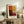 May include: Framed abstract art print with warm color blocks in red, orange, yellow, and burgundy tones displayed on rustic wooden sideboard. Modern minimalist poster in light wood frame sits alongside fern plant, ceramic vase, stacked books, and brown leather chair in neutral living room setting with white walls and natural lighting.