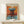May include: Framed abstract art poster in warm wood frame leaning against white wall on light wooden surface. Artwork features orange and blue rectangular forms with painterly brushstrokes in Rothko's signature style. Small white wrapped gift box with twine sits in front of frame.