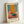 May include: Framed abstract art print in light wood frame leaning against white wall. The artwork features bold geometric blocks of orange, teal blue, and coral pink colors in Rothko's distinctive style. Artist name "MARK ROTHKO" printed below the colorful composition on cream background. Natural lighting creates soft shadows on the wall behind the vertically oriented rectangular frame.