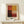 May include: Framed abstract art print in warm earth tones - deep reds, golden yellow, burgundy, and cream arranged in geometric color blocks. Print displays "MARK ROTHKO" text below artwork. Light wood frame with cream matting sits on pale wooden surface beside small white wrapped gift with evergreen sprig. Neutral beige wall background with soft natural lighting.