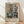 May include: Framed abstract art print leaning against white wall on light wooden floor. The artwork features geometric shapes in muted teal, orange, and beige tones depicting stylized kitchen objects like bowls and containers. Natural wood frame with white matting. Soft natural lighting creates diagonal shadows across the white textured wall.