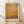 May include: Framed art print in light wood frame leaning against white wall on wooden floor. Artwork shows abstract painting with warm golden yellow and amber tones blending into darker brown at bottom, with soft cloudy textures throughout. White matting surrounds the image with artist name below in dark text. Natural window light creates diagonal shadows across the white wall behind the frame.