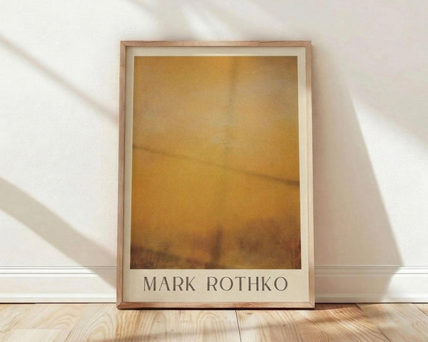 May include: Framed art print in light wood frame leaning against white wall on wooden floor. Artwork shows abstract painting with warm golden yellow and amber tones blending into darker brown at bottom, with soft cloudy textures throughout. White matting surrounds the image with artist name below in dark text. Natural window light creates diagonal shadows across the white wall behind the frame.