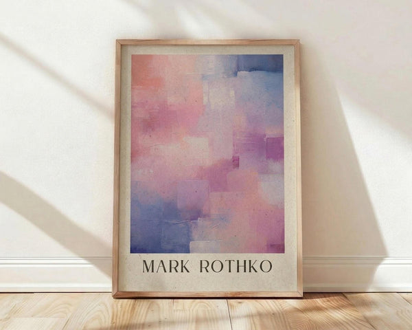 May include: Framed poster print featuring abstract watercolor artwork with soft pink, purple, and blue rectangular color blocks in gradient wash style. Light wood frame with cream matting displays "MARK ROTHKO" text below artwork. Print leans against white wall on natural wood floor with sunlight casting diagonal shadows across cream textured wall.
