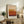 May include: Framed art print featuring horizontal color bands in cream, yellow, orange, and burgundy tones displayed on rustic wooden sideboard. Scene shows neutral living room with fern plant, stacked books, ceramic vase, and beige upholstered chair. Natural lighting creates soft shadows on white wall behind minimalist abstract artwork in light wood frame.