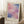 May include: Framed abstract watercolor art print in soft pink, purple, and blue tones leaning against white paneled wall. Light wooden frame with cream matting displays artist name at bottom. Cozy room setting with white coffee cup on wooden block, beige knit throw, and natural sunlight casting geometric shadows on white shag rug.