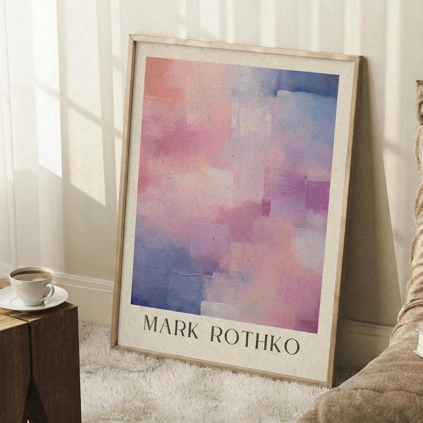 May include: Framed abstract watercolor art print in soft pink, purple, and blue tones leaning against white paneled wall. Light wooden frame with cream matting displays artist name at bottom. Cozy room setting with white coffee cup on wooden block, beige knit throw, and natural sunlight casting geometric shadows on white shag rug.