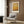 May include: Framed abstract art print with warm golden and amber tones hanging on wood-paneled wall above beige upholstered armchair with geometric patterned pillow. Modern reading nook with brass floor lamp, dark round side table with open book, and sheer curtains filtering natural light.