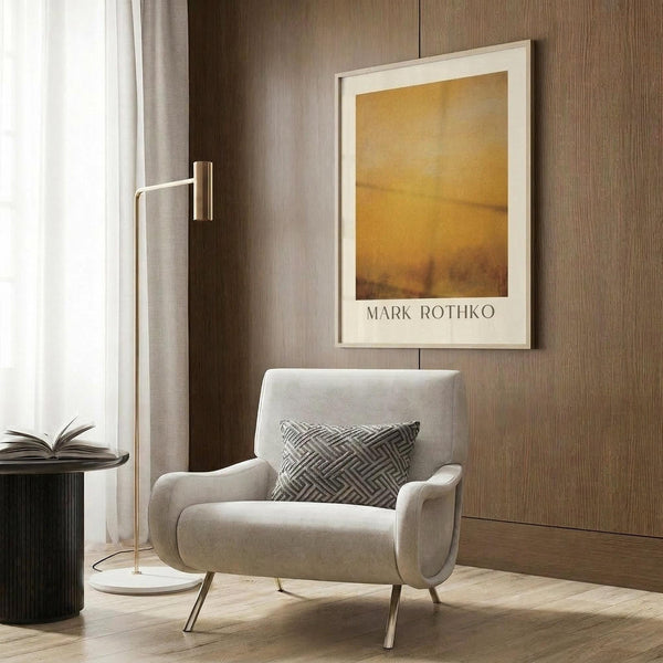 May include: Framed abstract art print with warm golden and amber tones hanging on wood-paneled wall above beige upholstered armchair with geometric patterned pillow. Modern reading nook with brass floor lamp, dark round side table with open book, and sheer curtains filtering natural light.