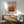 May include: Framed abstract art print with horizontal color bands in cream, yellow, orange, and burgundy tones displayed above a wooden sideboard. Mid-century modern living room setting with mint green upholstered chair, white ceramic table lamp, and natural sunlight casting shadows across white walls.