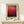 May include: Framed art print displaying abstract color field painting with gradient from light orange-pink at top transitioning to deep burgundy and dark brown at bottom, displayed in light wood frame with cream matting. Small white wrapped gift with evergreen sprig sits on light wooden surface below frame against cream wall.