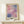 May include: Abstract watercolor artwork with gradient blocks in pink, purple, coral and blue tones displayed in light wood frame on cream wall. Small white gift box with green sprig sits on white wooden table beneath the framed print. Soft natural lighting creates gentle shadows on minimalist neutral background.