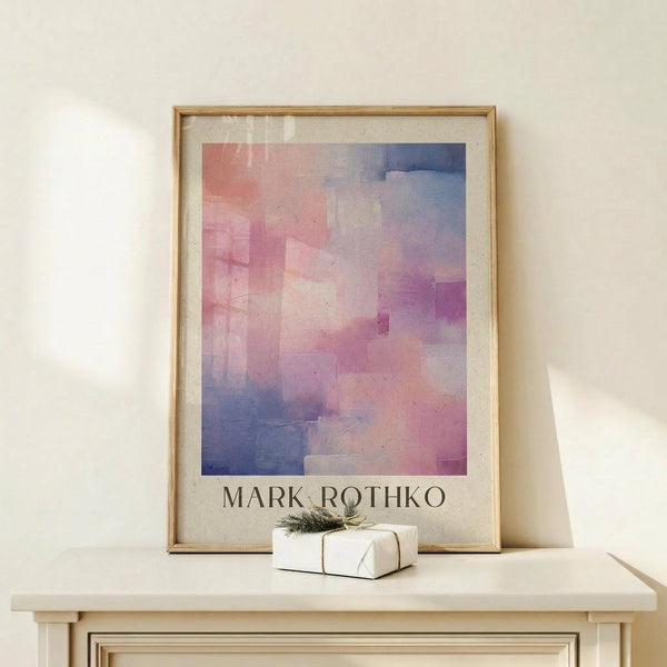 May include: Abstract watercolor artwork with gradient blocks in pink, purple, coral and blue tones displayed in light wood frame on cream wall. Small white gift box with green sprig sits on white wooden table beneath the framed print. Soft natural lighting creates gentle shadows on minimalist neutral background.