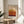 May include: Framed abstract art print with horizontal color bands in warm tones - cream, yellow, orange, and deep burgundy - displayed on a round wooden side table next to a beige upholstered chair. Sunlight streams across the neutral-toned room, casting shadows from a plant's leaves. The artwork features the artist's name below the geometric composition.