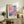 May include: Framed abstract art print with pink, purple and blue watercolor-like washes displayed on rustic wooden sideboard beside fern plant, ceramic vase, and stacked books in warm-toned living room setting with cream upholstered chair visible in foreground.