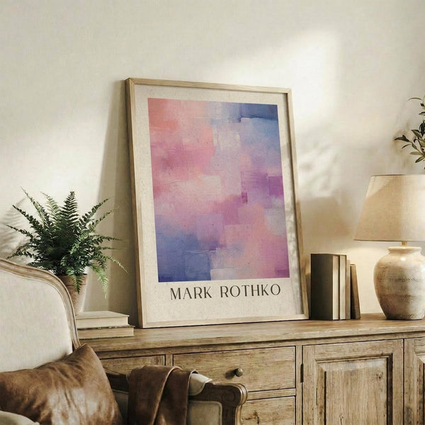 May include: Framed abstract art print with pink, purple and blue watercolor-like washes displayed on rustic wooden sideboard beside fern plant, ceramic vase, and stacked books in warm-toned living room setting with cream upholstered chair visible in foreground.