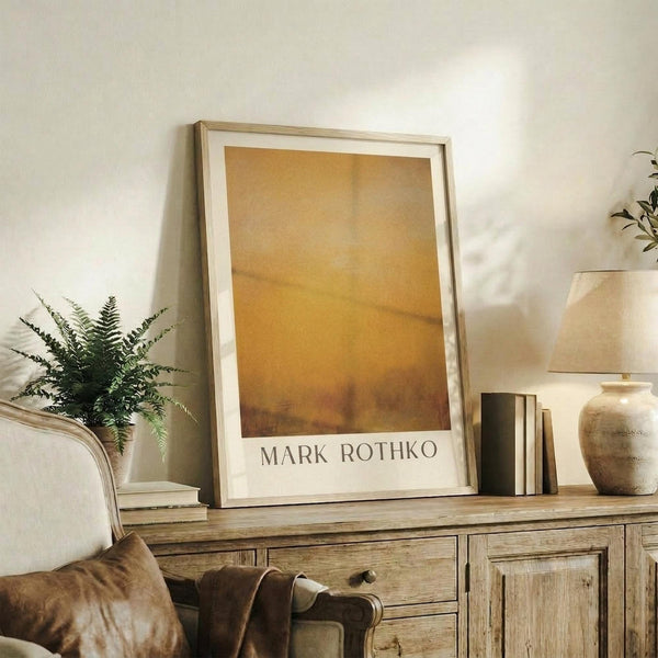 May include: Framed poster with warm golden orange abstract painting in light wood frame, displayed on rustic wooden sideboard alongside fern plant, ceramic vase, and books in neutral-toned living room with beige chair and white walls