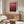 May include: Abstract art poster with red and burgundy gradient colors displayed above a wooden credenza in a modern living room. White sculptural chair and glass coffee table visible in foreground. Poster shows deep crimson tones fading to darker maroon, framed in light wood frame against neutral wall.