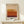 May include: Framed art print displaying horizontal color bands in warm earth tones - cream, tan, golden yellow, orange, rust, and deep burgundy - with "MARK ROTHKO" text below. Light wood frame with white matting, displayed on pale wooden surface alongside small white wrapped gift with green sprig. Soft natural lighting against cream wall.