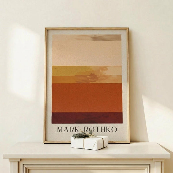 May include: Framed art print displaying horizontal color bands in warm earth tones - cream, tan, golden yellow, orange, rust, and deep burgundy - with "MARK ROTHKO" text below. Light wood frame with white matting, displayed on pale wooden surface alongside small white wrapped gift with green sprig. Soft natural lighting against cream wall.