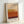 May include: Framed art print featuring horizontal bands of color transitioning from cream at top through yellow, orange, to deep burgundy at bottom, inspired by abstract expressionist style. Light wood frame with white matting displays "MARK ROTHKO" text below the artwork. Print leans against white wall corner, showing warm earth tone palette with soft, painterly edges between color blocks.
