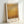 May include: Framed abstract painting print in warm golden and amber tones with soft, hazy brushstrokes creating a dreamy, atmospheric effect. The artwork features horizontal bands of color transitioning from lighter yellows at top to deeper oranges and browns at bottom. Displayed in a light wooden frame with white matting, leaning against a white wall in soft natural lighting.