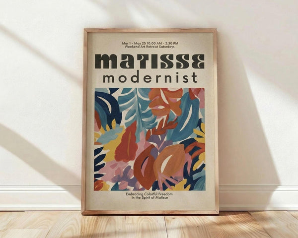 May include: Framed poster with cream background displaying abstract botanical artwork in vibrant blues, oranges, reds, and yellows. The colorful cut-out style design features flowing organic shapes and leaf-like forms. Bold black text reads "matisse" and "modernist" at top, with smaller text below the artwork. Natural wood frame leans against white wall on light wooden floor with soft window light creating shadows.