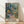 May include: Framed art print leaning against white wall on light wood floor. Colorful botanical illustration shows lush tropical garden scene with large coral and yellow flowers, palm fronds, and small figures on winding path. Vibrant blues, greens, oranges in layered collage style. "MATISSE" text at bottom in brown letters. Natural wood frame with warm lighting creating soft shadows on textured white wall.