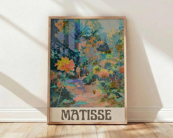 May include: Framed art print leaning against white wall on light wood floor. Colorful botanical illustration shows lush tropical garden scene with large coral and yellow flowers, palm fronds, and small figures on winding path. Vibrant blues, greens, oranges in layered collage style. "MATISSE" text at bottom in brown letters. Natural wood frame with warm lighting creating soft shadows on textured white wall.