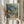 May include: Person holding framed art print featuring vibrant tropical garden scene with lush foliage in blues, greens, oranges and yellows, small figure on winding path, "MATISSE" text at bottom, natural wood frame, dried pampas grass beside frame, neutral beige clothing and curtained background
