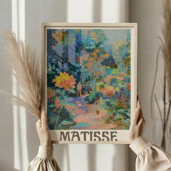 May include: Person holding framed art print featuring vibrant tropical garden scene with lush foliage in blues, greens, oranges and yellows, small figure on winding path, "MATISSE" text at bottom, natural wood frame, dried pampas grass beside frame, neutral beige clothing and curtained background