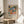 May include: Framed poster with colorful botanical artwork featuring tropical leaves, fruits, and plants in vibrant blues, oranges, greens, and pinks displayed on a round wooden side table next to a beige upholstered chair with decorative pillow, palm plant visible in corner, natural sunlight streaming across neutral wall creating soft shadows