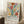 May include: A framed poster with colorful abstract floral design in Matisse-inspired style, featuring bold shapes in red, yellow, green, orange, and purple on cream background. The artwork shows stylized flowers, leaves, and organic forms with "Celebrating Colors of Life" text at bottom. Displayed in light wood frame leaning against white wall on textured rug, with coffee cup on wooden stool nearby in bright, airy room setting.