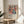 May include: Framed colorful poster featuring abstract woman figure with flowers in Matisse-inspired style, displayed on wooden round side table next to beige upholstered chair. Artwork shows vibrant blues, corals, greens in cut-out aesthetic with "Celebrating Colors of Life" text below image. Natural lighting creates soft shadows across neutral wall background with plant leaves visible.
