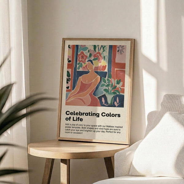 May include: Framed colorful poster featuring abstract woman figure with flowers in Matisse-inspired style, displayed on wooden round side table next to beige upholstered chair. Artwork shows vibrant blues, corals, greens in cut-out aesthetic with "Celebrating Colors of Life" text below image. Natural lighting creates soft shadows across neutral wall background with plant leaves visible.