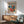 May include: Framed modernist poster with colorful abstract leaf and floral shapes in blues, oranges, reds, and yellows, displayed above a wooden sideboard with white ceramic lamp. Light green upholstered armchair with wooden frame sits nearby on wooden floor with natural sunlight casting shadows on white wall.