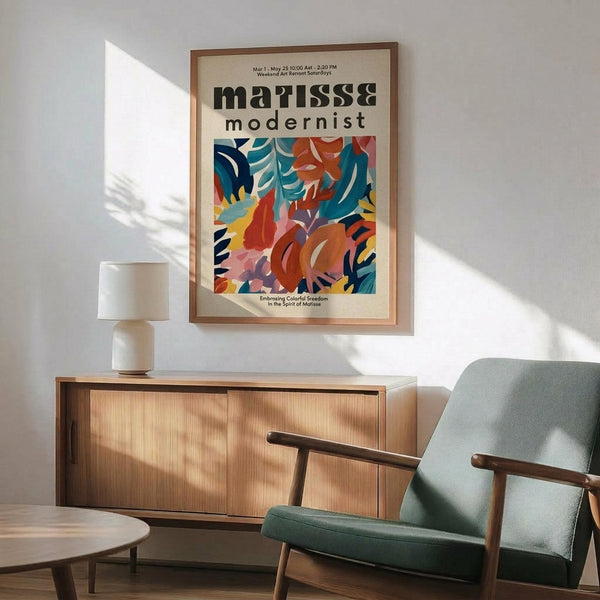 May include: Framed modernist poster with colorful abstract leaf and floral shapes in blues, oranges, reds, and yellows, displayed above a wooden sideboard with white ceramic lamp. Light green upholstered armchair with wooden frame sits nearby on wooden floor with natural sunlight casting shadows on white wall.