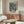 May include: Colorful botanical art print with "MATISSE" text displayed above wooden sideboard in modern living room. Vibrant tropical garden scene features blues, greens, oranges in painterly style. Thin wooden frame complements walnut furniture, white sculptural lamp, cream chair, and glass coffee table on neutral walls.