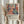 May include: Person in cream ruffled sleeves holding framed poster featuring abstract woman in coral dress surrounded by colorful flowers and foliage in Matisse-inspired style. Light wooden frame against neutral curtained background with dried wheat stems visible. Bold geometric shapes in coral, blue, green, and pink tones create vibrant artistic composition.