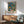 May include: Framed art print featuring vibrant tropical garden scene with lush foliage in blues, greens, yellows and coral tones, displayed above mid-century modern wooden credenza with ceramic table lamp. Mint green upholstered armchair with wooden frame sits nearby on light wood floors. Warm natural sunlight creates geometric shadows on white wall, showcasing the colorful botanical artwork in wooden frame.