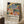 May include: Framed poster with colorful Matisse-style botanical illustration featuring tropical leaves, fruits, and abstract shapes in blues, greens, oranges, and pinks on cream background. White text below reads "Celebrating Colors of Life" with descriptive paragraph. Light wood frame leans against white wall corner next to coffee cup on wooden side table and cream knit throw blanket on white textured rug.