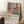May include: Framed colorful abstract poster leaning against white wall beside cozy armchair and wooden side table with coffee cup. Artwork features stylized woman figure surrounded by vibrant flowers and foliage in Matisse-inspired cut-out style using coral, blue, green and pink tones. Bold text reads "Celebrating Colors of Life" with descriptive text below. Natural lighting creates soft shadows on textured rug.