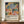 May include: Framed poster with colorful Matisse-style artwork featuring abstract botanical elements including a blue tree, tropical leaves, fruits, and organic shapes in vibrant blues, greens, oranges, and pinks on cream background. Text below reads "Celebrating Colors of Life" with descriptive paragraph. Displayed on wooden sideboard next to white ceramic vase and stack of books against cream wainscoted wall.