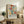 May include: Colorful abstract floral poster with vibrant red, yellow, teal and pink organic shapes in Matisse cut-out style, displayed in light wood frame on rustic wooden sideboard. Room setting shows cream upholstered chair, small potted fern, ceramic vases, and stacked books against white wall with warm natural lighting.
